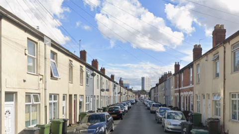 Terraced houses can be seen either side of the street with cars parked outside. The houses are mainly painted cream, with a few in grey and plain brick. A block of flats can be seen far off in the distance.