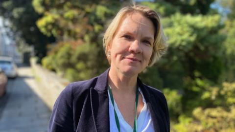 A gently-smiling woman with blonde, shoulder length hair in a white blouse and navy blue blazer, with a green lanyard on. She stands on a pavement with bushes and cars behind her on a sunny day.