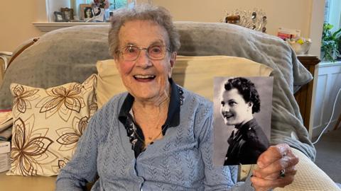 Sheila Pantin has short grey hair and is wearing glasses, a blue cardigan over a black top. In her left hand she is holding a black and white photograph her herself in military uniform