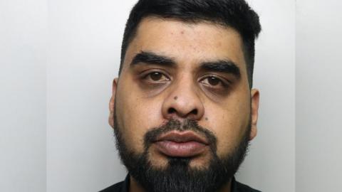 A police custody picture of a man staring straight ahead. He has black hair and a black beard and is wearing a black top. 