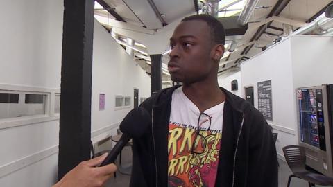 A young black man talking into a microphone in a college corridor. He is wearing a black hoodie and white t-shirt with an orange and red drawing