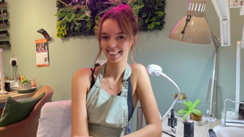A young woman with auburn hair tied up smiles at the camera from her workstation inside the nail salon. She wears a light green apron over a black top and has a smartwatch on her wrist. Behind her, a pink neon sign reading 'Taylor Made' glows against a wall of artificial greenery, with nail lamps and bottles of polish on the desk beside her.
