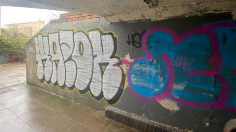 A large colourful piece of graffiti on a wall in a crumbling underpass. It is a wet day and the pavement has water on it.