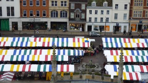 Aerial photo of Cambridge's Market Square. The stalls have canopys which are white and stripped colours and are in three long rows. Behind them are a row of shop.