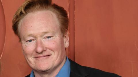 Conan O'Brien, in a blue shirt and dark suit coat, stands in front of an orange backdrop as he attends the HBO & Max Post-Emmy Reception at San Vicente Bungalows