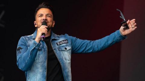 Peter Andre wearing a black t-shirt and denim jacket, performing on a stage.