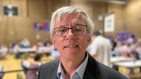 Steve Jarvis with short white hair and glasses, looking at the camera while wearing a dark grey jacket and a light grey shirt. There is an election count going on behind him in a sports hall.