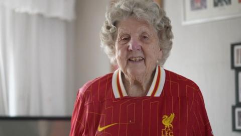 Peggy Pickavant is smiling at the camera. She is wearing a Liverpool FC shirt and is standing in her living room in front of a TV with photographs hanging on the walls.