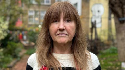 Louise Gates in black and cream striped jumper. She has brown hair past her shoulders and a fringe. She stands in the community garden, which is blurred behind her. 