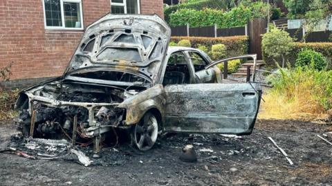 A burnt out shell of a car which was on fire. Debris and charred materials can be seen surrounding the vehicle.