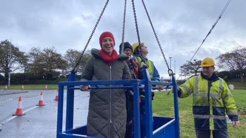A woman and a man in a crane surrounded by two men in high vis and hard hats on a road.