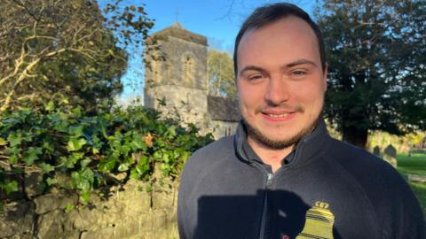 A young man in blue fleece smiling with a church, foliage and some graves in the background.