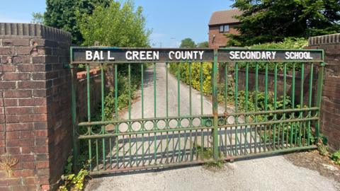 Between two red and brown brick pillars is a metal green gate that has rusted. Across the top it says "Ball Green County Secondary School" in white writing. beyond the gate is a concrete path with trees and bushes either side, as well as walls on either side. A house sits in the background behind a large tree