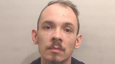 A balding brown haired man has a short brown moustache and short brown beard.