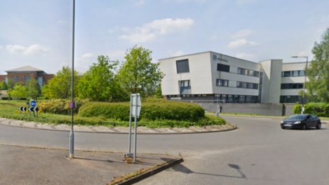 Two cars travelling around the roundabout on Colney Lane with the Norfolk and Norwich University Hospital in the background.