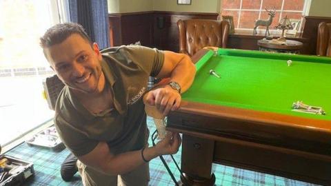 A man learn over next to a snooker table and smiles at the camera. He wears a dark green polo shirt which says 'AC Snooker' on it.