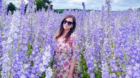 A woman wearing a white and pink floral dress middle of a flower field. Dozens of blooming lilac and purple flowers are around her.