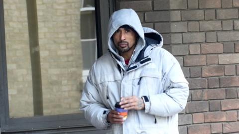 A man wearing a grey hoodie and a grey jacket. He is holding a coffee cup.