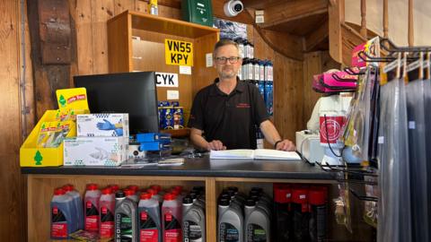 Martin Gillott stands behind his shop till. Around him is car equipment: number plates, windscreen wash and car air fresheners