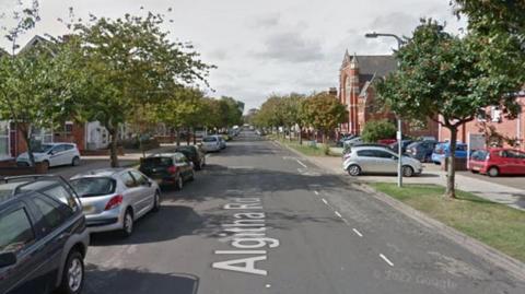 A long, straight and wide road is flanked by trees and houses. Cars are parked on the side of the road.