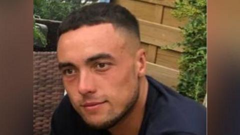 Kieran Shepherd sitting outside wearing a navy shirt. He has black hair and has a faint smile in the photo.