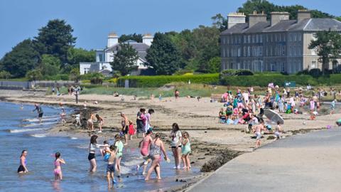 Crowds of people on a beach and in the sea. A large house sits on the shoreline.
