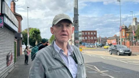 James Smith, wearing a grey baseball cap and a grey zip-up jacket, stands on a busy road lined by shops, in the background a blue, red and white Union Jack flag attached to a lampost is fluttering in the breeze