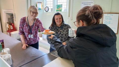 Two women volunteers - one wearing a purple floral top - handing out a plate of toast to a lady in a black jacket, who is also holding a cup of tea and has her back to the camera. Another volunteer is wearing an animal-print hoodie smiling at the lady receiving the toast.