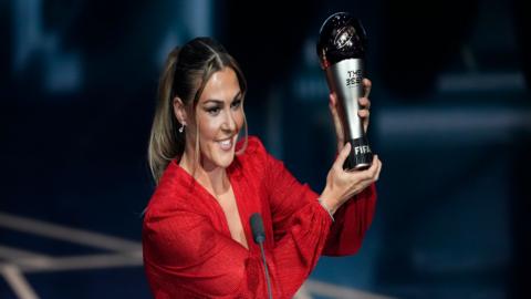 Mary Earps holds up a trophy. She is wearing a red dress and is smiling. She is stood on a stage and has brown hair that is tied into a ponytail.