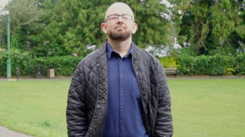 A bald and beared bespectacled man in his thirties, wearing a blue shirt and trousers and a dark jacket, stands in a park with his hands in his pockets.
