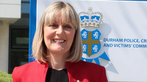 Joy Allen standing in front of a blue and white Durham Police sign. She has a blonde, collar-length bob with a fringe and is wearing a red blazer with black top.