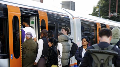 Passengers board a crowded London Overground train, with people queuing at the open doors of an orange-and-white carriage.