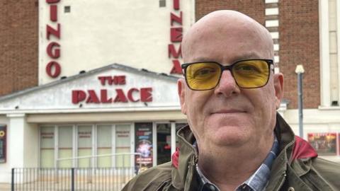 Mark Jones stands in front of the cinema. He is bald and wears glasses with yellow frames as well as a green coat and blue chequered shirt.
