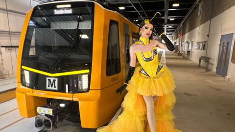 Sally TM posing to the right of one of the new dark yellow Metro trains. She has short, brown hair which is curled underneath, with a streak of yellow dye at the front. A piece of yellow hair is standing up above her head. She is wearing a yellow dress, which is lighter than the train, which is made up of a corset with silver accents and a tulle skirt which is short at the front and long at the back. She is also wearing a yellow collar around her neck and black long gloves.