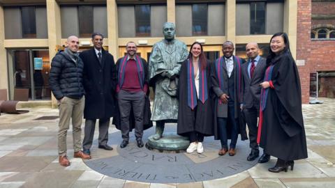 A posed group shot featuring students who took part in the specialist kidney training, together with consultants who created the court. They are five men and two women lining up either side of a metal statue outside Newcastle University.
Four of them, two men and two women, wear a graduation gown with a blue and red and blue stole. The other three men are dressed in formal wear.