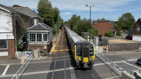 A South Western Railway train crossing a level crossing, where gates are down stopping cars from crossing the road.