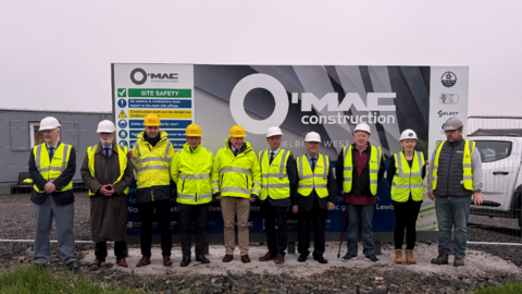 Ten representatives from SSEN Transmission and Hebridean Housing Partnership in hi-vis jackets and helmets, standing in front of a black and white construction billboard. Grey skies behind. Fencing, grey portacabin and white construction vehicle in the background. 