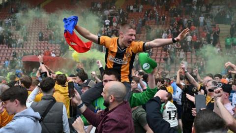 Fans celebrating inside the stadium - with one fan in a yellow and black jersey sitting on the shoulder of another fan. 