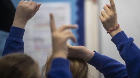 A stock image of children raising their hands in a classroom setting. The children are pictured from behind and the focus is on their raised hands. They are wearing blue uniform jumpers. The background is blurred.