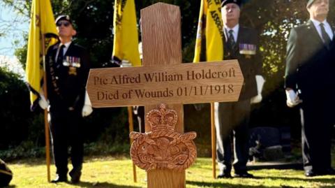 A close-up of a wooden cross which says "Pte Alfred William Holdcroft, died of wounds 01/11/1918". It has a crest underneath the words. Four men are standing behind it on the grass wearing uniforms and holding large yellow flags.