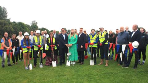 A group of people stand on  grass among red, white and blue bunting. Some of the people are holding shovels and there are trees visible in the background.