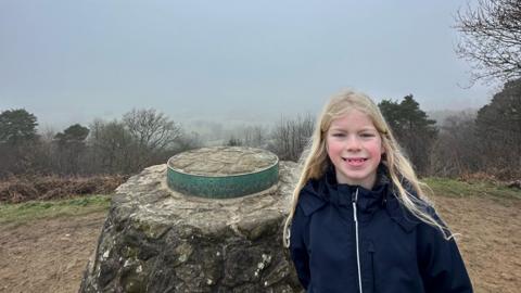 A boy pictured at the top of the Surrey Hills. he has long blonde hair and is wearing a navy raincoat. It is foggy and little can be seen in the background. 