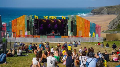 A colourful outdoor stage labelled 'THE VIEW' set up on a grassy area overlooking a beach and the ocean. The stage features vibrant multi-coloured panels and purple lighting. A small crowd sits on the grass watching a performance, with cliffs and the shoreline visible in the background.