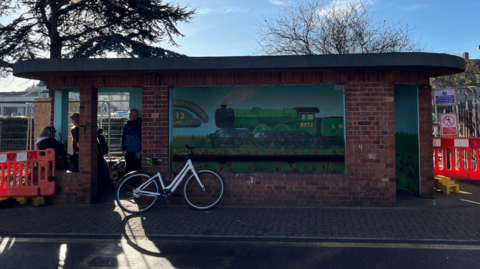 Sheringham bus shelter. It is a brick building with a flat roof built in an art deco style. There is a mural of a train on the inside. There is a bicycle leaning against the front of the shelter. There are three people inside. They are gathered on the left hand side. Behind and beside the shelter there is wire fencing and orange-and-white barriers.
