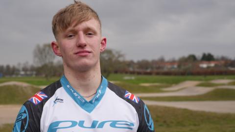 Jared Hill stands in front of a BMX track. He is wearing a V-necked, white, black and sky blue racing jersey with Union flags on each shoulder and the word "Pure" across the chest.