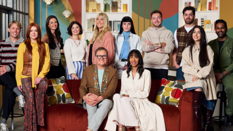 Alan Carr is sat down on a red settee wearing a brown jacket and grey trousers. He is sat next to judge Michelle Ogundehin, who is dressed in a cream jacket and matching skirt. They are joined by ten people, who are all contestants on the series Interior Design Masters. Sophie from Stoke on Trent is among the contestants and is second left in the photograph. She has long red hair and is wearing a yellow shirt, patterned red trousers and a brown gilet. Ben, from Kinver, is stood behind the red sofa with his arms folded, wearing a beige hoodie.