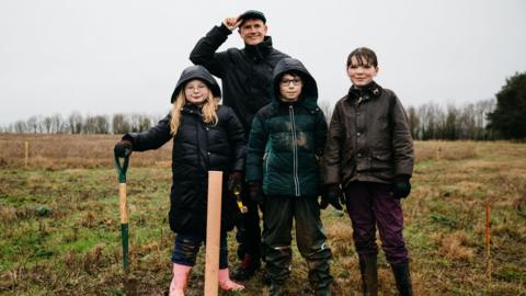 Luke stands in a black jacket and black flat cap, smiling at the camera. There are three smiling children in front of him, all in winter coats, one of whom is holding a spade. There is a grassy open space behind them, and the sky is grey. 