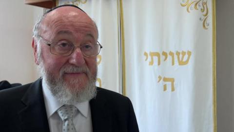 The Chief Rabbi wears a dark suit, white shirt, grey tie with a darker grey square pattern, and a kippah. He wears glasses and has a white beard.