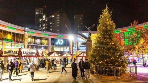 The centre of Woking with shops seen in the background including JD Sports and Pret a Manger. It is night time and a Christmas tree in the foreground is lit up and there is also a sausage stand and Christmas market stalls