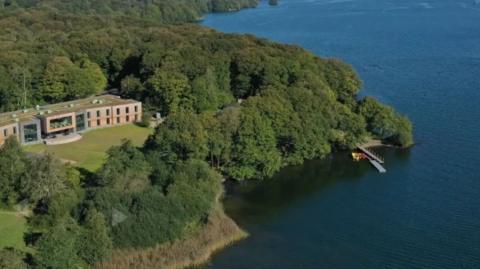 An aerial view of the YMCA Lakeside camp in Windermere, which is next to the lake. It has a modern building with a big box window next to it and is surrounded by trees. There is a jetty.